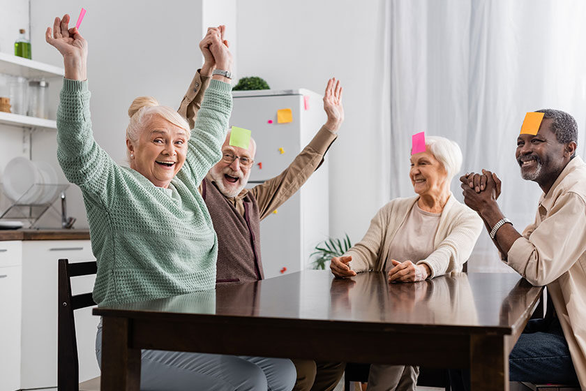 Excited and multicultural senior friends with sticky notes on foreheads playing game in kitchen